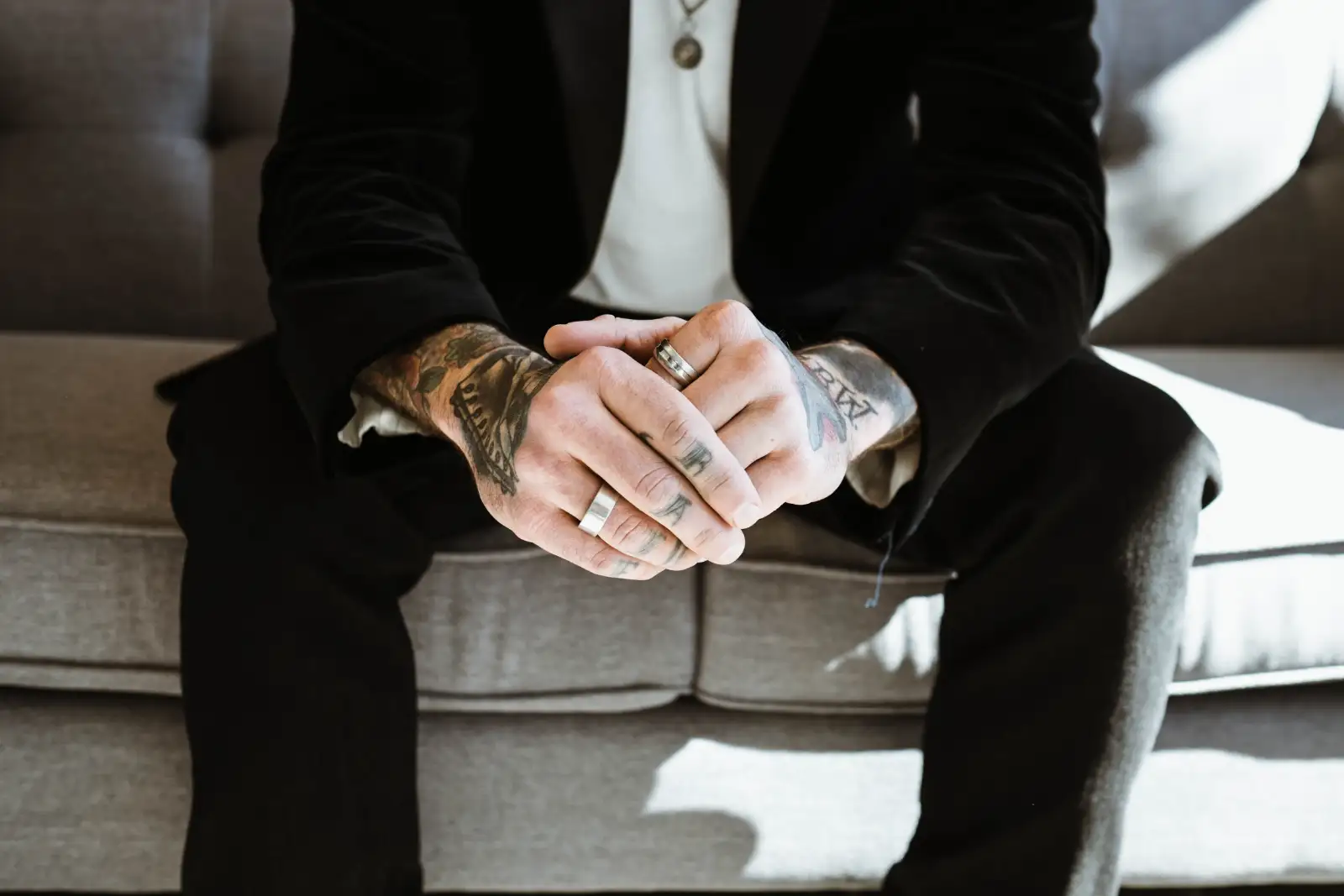 Tattooed hands with silver rings, clasped over black trousers